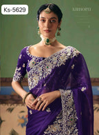 A woman models a purple georgette saree with intricate silver and gold floral embroidery and green stone accents, paired with matching jewelry. The background is a soft olive green wall with a patterned rug visible at the bottom.